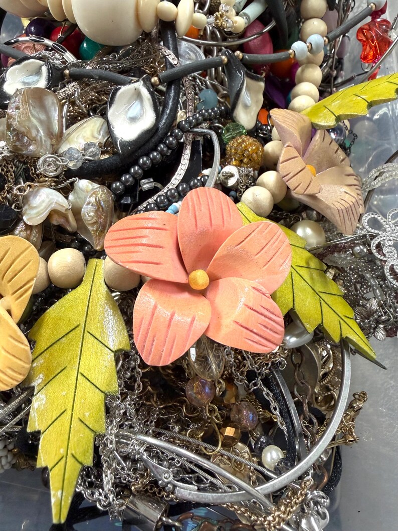 May include: A jumble of jewellery items, including necklaces, bracelets, and earrings. The collection features silver chains, beaded strands, and pendants. Wooden flower and leaf accents in peach and yellow add colour and natural elements to the mix.