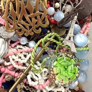 May include: A collection of various jewelry pieces, including necklaces, pendants, and charms. The image features a gold-colored monogram pendant, a miniature Eiffel Tower charm, and a glass orb pendant with a green floral design. Beads in various colors are also visible.