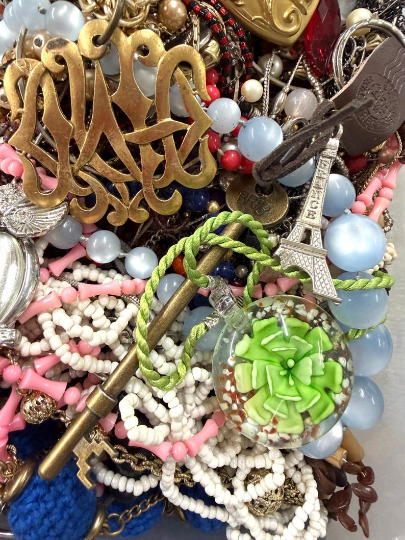 May include: A collection of various jewelry pieces, including necklaces, pendants, and charms. The image features a gold-colored monogram pendant, a miniature Eiffel Tower charm, and a glass orb pendant with a green floral design. Beads in various colors are also visible.