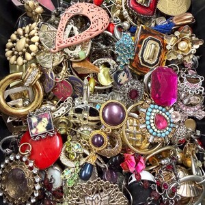 May include: A collection of vintage jewelry, including earrings, brooches, and pendants. The pieces feature various colors, shapes, and materials, such as gold, silver, red, pink, and turquoise. Some items have gemstones and intricate designs.