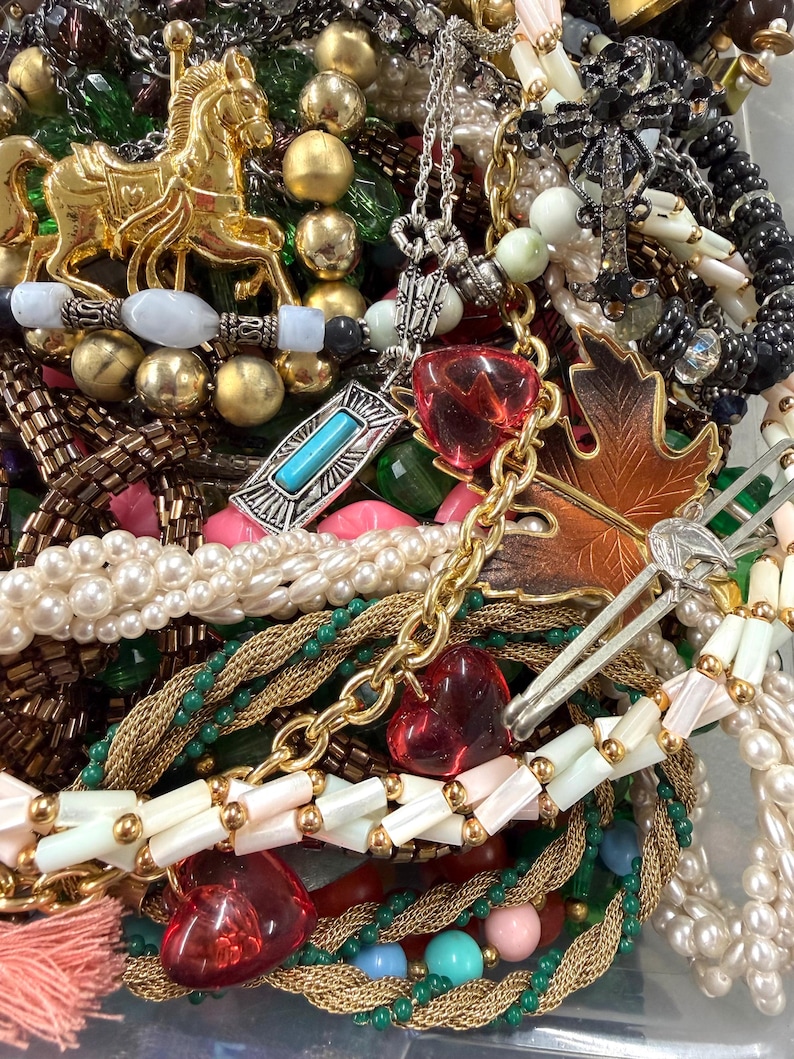 May include: A collection of vintage jewelry including necklaces, bracelets, and pendants. The assortment features gold-toned metal, faux pearls, and colorful beads. Notable pieces include a gold carousel horse pendant and heart-shaped charms.