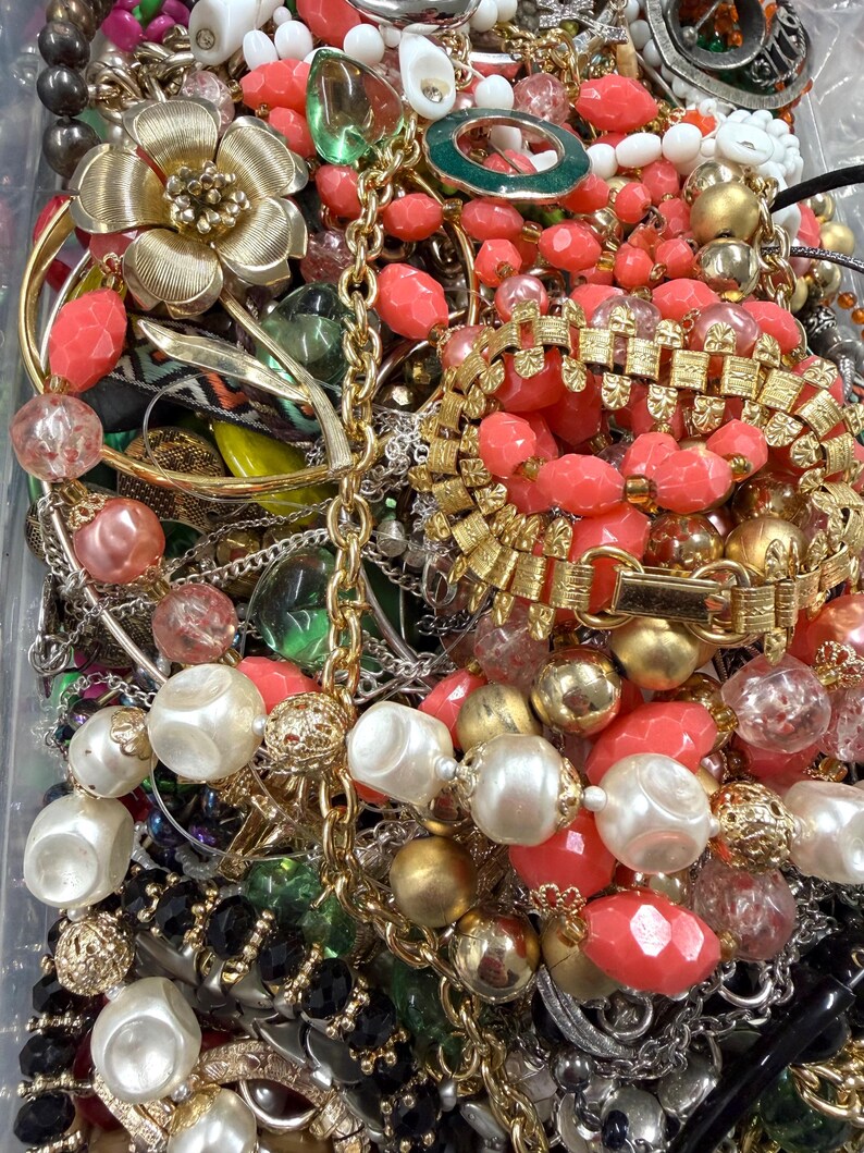 May include: A collection of vintage jewelry including necklaces, bracelets, and earrings. The pieces feature a variety of colors, including gold, coral, white, and black. Some items have floral and geometric designs, with pearls and beads.