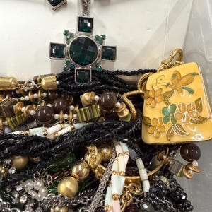May include: A collection of jewelry including necklaces, earrings, and bracelets. The necklaces feature a variety of beads, chains, and pendants. One pendant is a cross shape with green stones. Another is a yellow rectangular pendant with a butterfly and floral design.