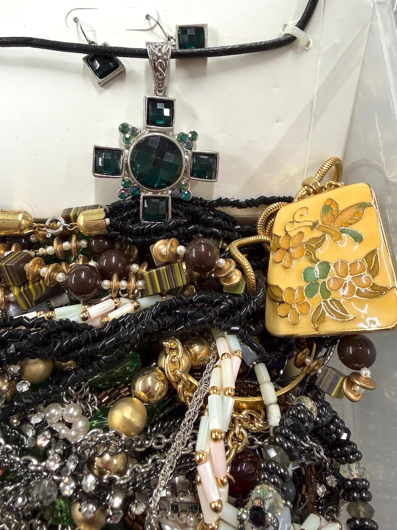 May include: A collection of jewelry including necklaces, earrings, and bracelets. The necklaces feature a variety of beads, chains, and pendants. One pendant is a cross shape with green stones. Another is a yellow rectangular pendant with a butterfly and floral design.