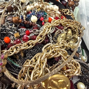 May include: A close-up shot of a collection of assorted jewelry, including necklaces, bracelets, and beads. The jewelry features a variety of colors, including gold, silver, red, and black. There are chunky gold chains, pearl necklaces, and beaded bracelets.