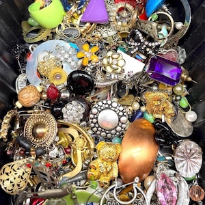 May include: A collection of vintage jewelry, including rings, brooches, earrings, and pendants. The assortment features various colors, shapes, and materials, such as gold, silver, pearls, and gemstones. A purple triangular object and a gold teddy bear pendant are visible.