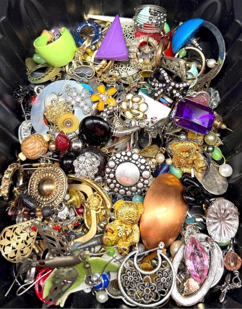 May include: A collection of vintage jewelry, including rings, brooches, earrings, and pendants. The assortment features various colors, shapes, and materials, such as gold, silver, pearls, and gemstones. A purple triangular object and a gold teddy bear pendant are visible.