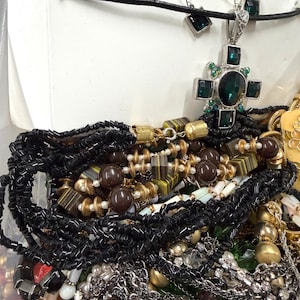 May include: A collection of various necklaces and jewelry. Includes a silver cross pendant with a large green stone and smaller green accents, black beaded necklaces, and other beaded and chain necklaces in gold, brown, and white.