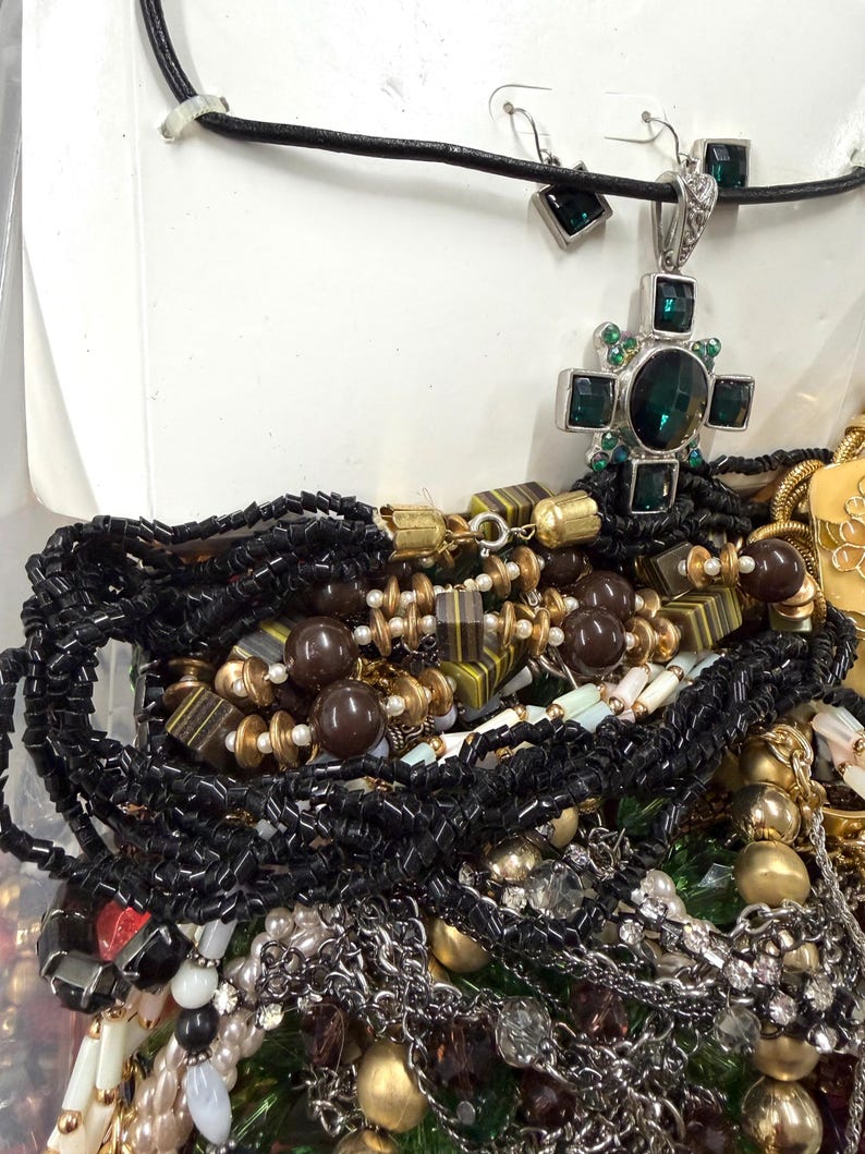 May include: A collection of various necklaces and jewelry. Includes a silver cross pendant with a large green stone and smaller green accents, black beaded necklaces, and other beaded and chain necklaces in gold, brown, and white.