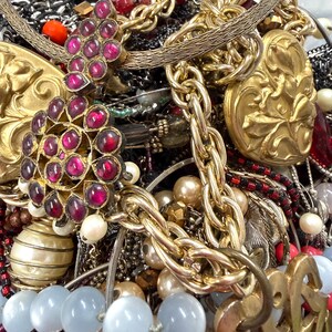 May include: A close-up of a collection of vintage jewelry. The image features gold chains, beaded necklaces, and pendants with red and pink gemstones. There are also pearl and blue-toned bead necklaces.