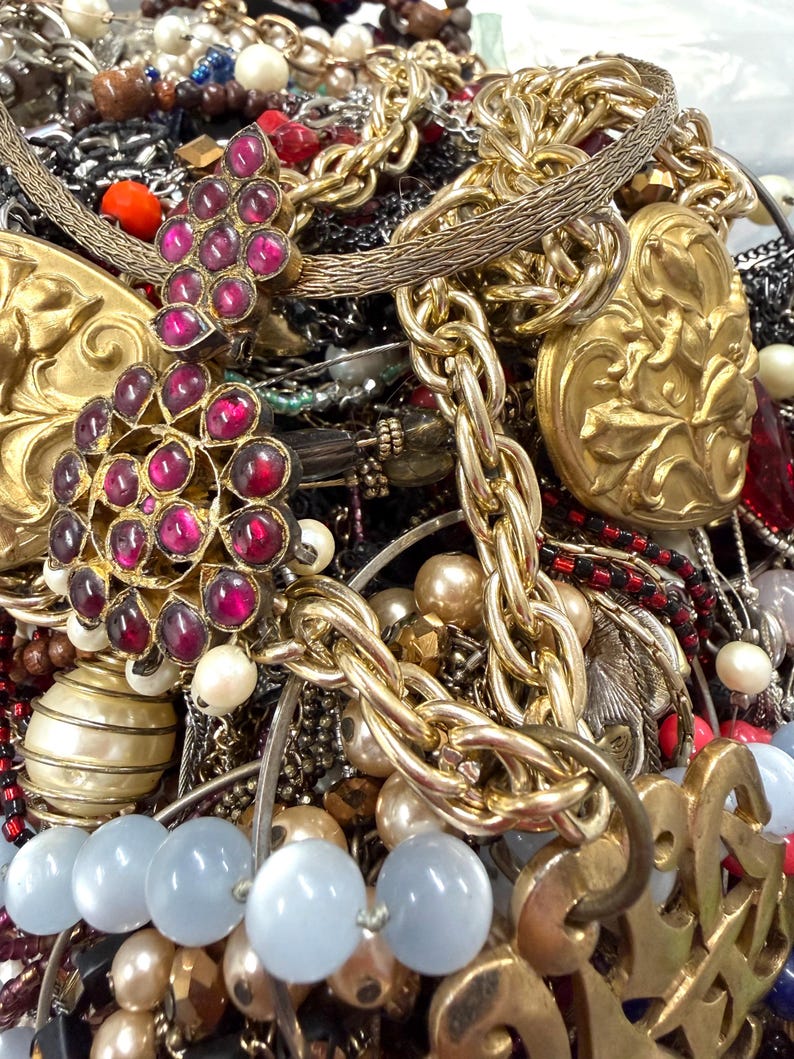May include: A close-up of a collection of vintage jewelry. The image features gold chains, beaded necklaces, and pendants with red and pink gemstones. There are also pearl and blue-toned bead necklaces.