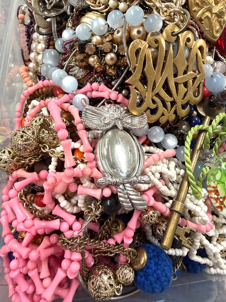 May include: A collection of vintage jewelry including necklaces, bracelets, and brooches. The assortment features pink coral beads, pearl strands, and a silver owl pendant. Gold-toned metal accents and blue beads add contrast to the mix.