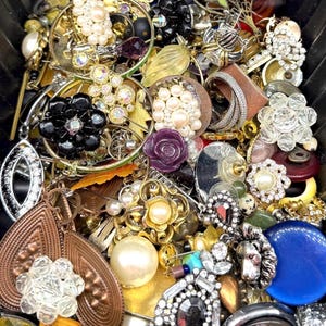 May include: A collection of vintage jewelry, including earrings, brooches, and rings. The pieces feature various designs, including pearls, rhinestones, and floral motifs. Colors include gold, silver, black, and blue. The jewelry is arranged in a black container.