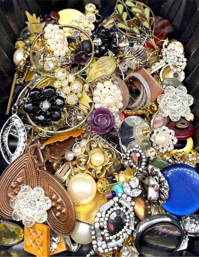 May include: A collection of vintage jewelry, including earrings, brooches, and rings. The pieces feature various designs, including pearls, rhinestones, and floral motifs. Colors include gold, silver, black, and blue. The jewelry is arranged in a black container.