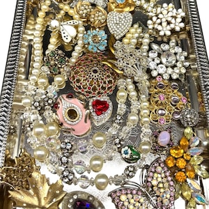 Puede incluir: Una bandeja plateada llena de una variedad de joyas vintage. La colección incluye collares de perlas, broches y piezas con incrustaciones de pedrería en varias formas y colores, incluyendo mariposas, corazones y diseños florales.