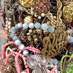 May include: A close-up view of a collection of vintage jewelry, including necklaces, bracelets, and pendants. The jewelry features various colors, including gold, silver, pink, blue, and red. Notable pieces include an owl pendant, beaded necklaces, and ornate brooches.