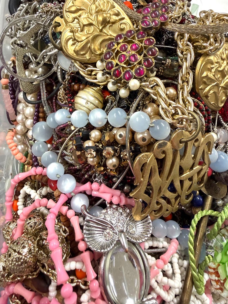 May include: A close-up view of a collection of vintage jewelry, including necklaces, bracelets, and pendants. The jewelry features various colors, including gold, silver, pink, blue, and red. Notable pieces include an owl pendant, beaded necklaces, and ornate brooches.