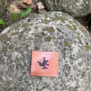 May include: A small, square, copper-colored tile with a black silhouette of a frog jumping. The tile has a slightly rough texture.