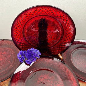 May include: Four red glass plates with a diamond pattern and a starburst design in the centre. The plates are stacked on top of each other.