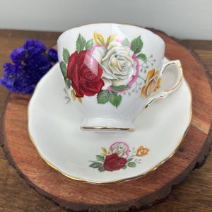 May include: A white teacup and saucer with a floral design featuring red, white, and yellow roses. The cup and saucer have a gold trim.