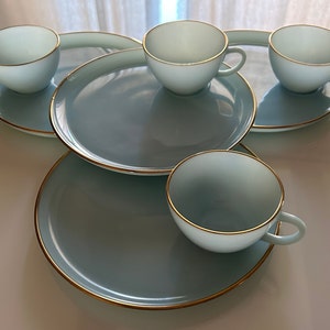 May include: Set of four light blue teacups and saucers with gold trim. The cups and saucers are arranged in a circle on a white surface.
