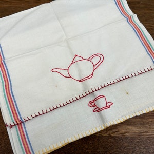 May include: A white tea towel with red embroidered images of a teapot and a cup. The towel has a multi-colored border with red, orange, yellow, blue, and green stripes.