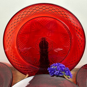 Puede incluir: Juego de cuatro platos de cena de vidrio rojo con un patrón de diamante y un diseño de estrella en el centro. Los platos están apilados sobre una superficie de madera con un pequeño ramo de flores moradas en frente.