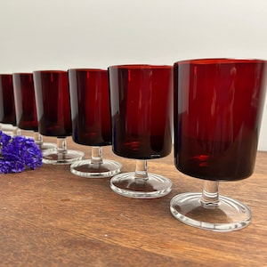 Puede incluir: Un juego de ocho copas de vino tinto con tallos y bases transparentes. Las copas están dispuestas en fila sobre una superficie de madera.