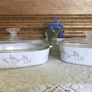 May include: Two white oval baking dishes with glass lids. The dishes have a floral design with purple irises and green leaves.