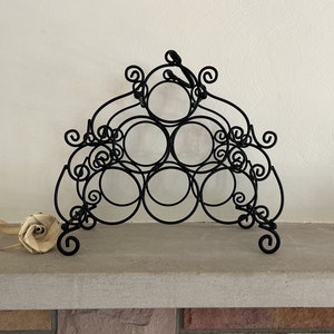 Puede incluir: Estante para vino de metal negro con un diseño decorativo en espiral. El estante tiene seis espacios circulares para guardar botellas de vino.