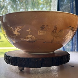Puede incluir: Un tazón de mezcla de cerámica marrón con un gallo dorado, un águila y otros diseños. El tazón está sentado en un soporte de madera con tres patas.