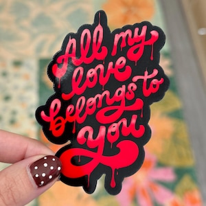 May include: A black sticker with the phrase "All my love belongs to you" in red, dripping font. The sticker has a unique shape and is held by a hand with a brown and white polka dot nail design.