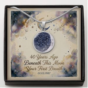 May include: A silver-toned circular pendant necklace featuring a dark blue star map design. The pendant rests on a card with a watercolour night sky background, a crescent moon, and the text 40 Years Ago Beneath This Moon Your First Breath 01.05.1987.
