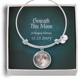 May include: A silver charm bracelet with a moon charm. The bracelet is in a white box with a blue background. The box has the text "Beneath This Moon" and "A Magical Moment 12.22.2003" on it.
