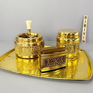 Pode incluir: Uma bandeja de metal dourado com acabamento martelado contém um conjunto de três acessórios de metal dourado correspondentes. O conjunto inclui um cinzeiro para cigarros com uma tampa de plástico branco, uma pequena caixa retangular e um frasco redondo com tampa. A bandeja está sobre um fundo cinza.