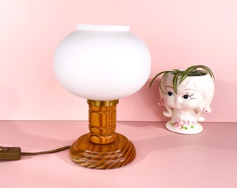 Puede incluir: Una pantalla de lámpara de vidrio blanco con una base de madera. La base es de madera marrón con un patrón geométrico. La lámpara está sobre una superficie rosa.