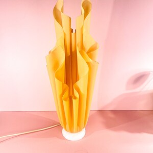 May include: A yellow, pleated lamp with a white base. The lamp is shaped like a flower and has a modern design.