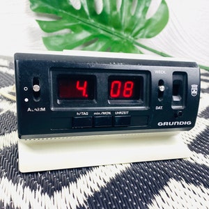 Pode incluir: Um relógio digital preto com um visor vermelho mostrando a hora 4:08. O relógio tem botões para alarme, hora, minuto e data. O relógio é etiquetado como "GRUNDIG" e tem uma base branca.