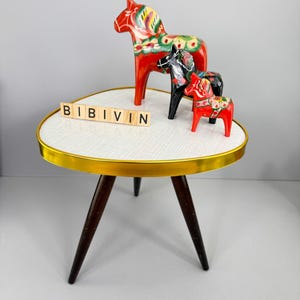 May include: Three Dala horses in red, black, and red, with floral patterns, stand on a small, white-topped table with a gold rim and dark wooden legs. The word "BIBIVIN" is spelled out in wooden letter tiles.