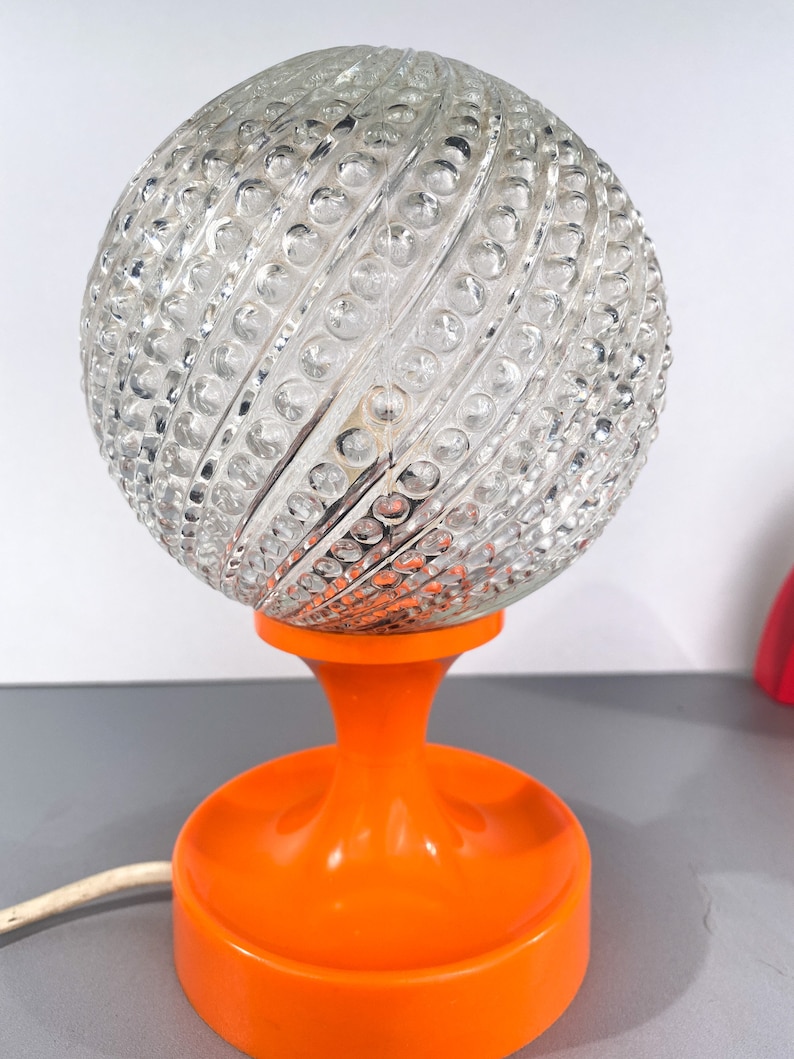 May include: A vintage orange table lamp with a clear glass globe shade. The globe has a textured, swirling pattern.