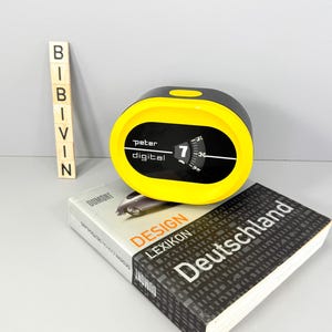 Könnte beinhalten: Eine schwarz-gelbe Digitaluhr mit dem Text "Peter digital" und der angezeigten Zahl "7". Die Uhr liegt auf einem Buch mit dem Titel "Design Lexikon Deutschland" von Dumont.