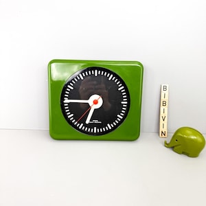 May include: A square, green clock with a black face and white markings. The clock has white hands and a red second hand. A small green elephant figurine and wooden letter blocks spelling "BIRVIN" are next to the clock.