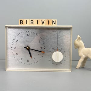 Könnte beinhalten: Eine cremefarbene Vintage-Uhr mit silbernem Zifferblatt und schwarzen Zeigern, mit einem Minutenzifferblatt. Rechts steht eine kleine Keramik-Hirschfigur. Das Wort "BIBIVIN" ist auf Holzblöcken über der Uhr geschrieben.