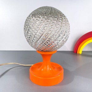 May include: A vintage table lamp with a clear glass globe shade featuring a textured, swirling pattern. The lamp base is a bright orange plastic with a round, wide base.