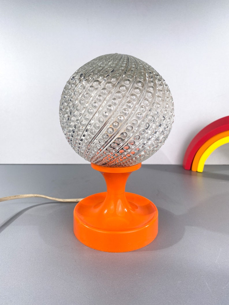 May include: A vintage table lamp with a clear glass globe shade featuring a textured, swirling pattern. The lamp base is a bright orange plastic with a round, wide base.
