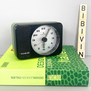 May include: A black and green alarm clock with a white face and black hands. The clock has a repeat button and the words "JUNGHANS" and "QUARTZ" printed on the face. The clock is sitting on top of a green and yellow book with the words "DESIGNLEXIKON ITALIEN" and "DUMONT" printed on the cover.