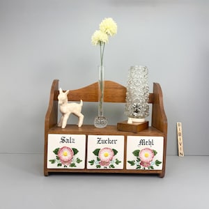 Puede incluir: Un especiero de madera con tres cajones de baldosas de cerámica etiquetados "Salz", "Zucker" y "Mehl". Los cajones presentan diseños florales. Una pequeña figura de ciervo, un jarrón de cristal con flores y una lámpara de cristal decorativa están en el estante.