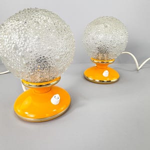 Könnte beinhalten: Zwei Vintage-Tischlampen. Jede Lampe hat eine klare, strukturierte Glaskugel auf einem leuchtend orangefarbenen Sockel mit goldenem Rand. Der Sockel hat einen weißen Ein-/Ausschalter. Die Lampen sind mit einem weißen Netzkabel verbunden.