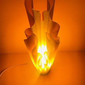 May include: A white, abstract, sculptural lamp with a warm, yellow light emanating from the center. The lamp is shaped like a flame and has a modern, minimalist design.