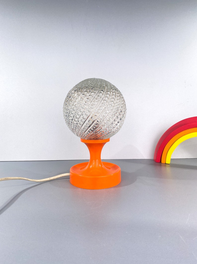 May include: A vintage table lamp with a clear glass globe shade with a textured pattern and an orange plastic base.